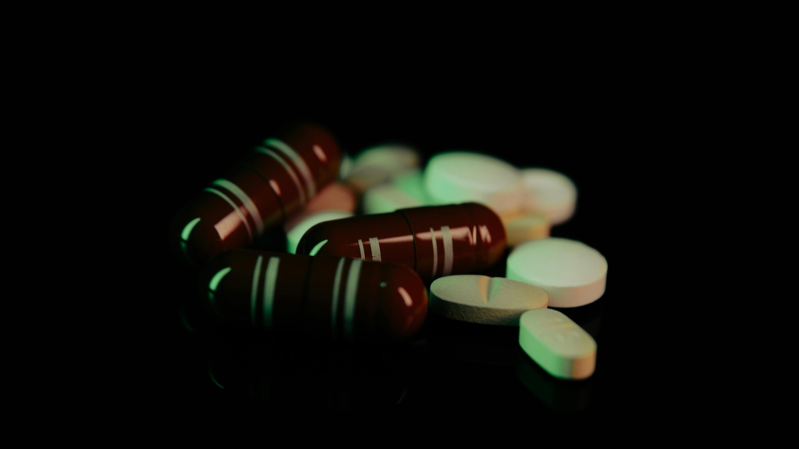 Assorted pills and capsules in green light on dark surface, representing pharmaceuticals.